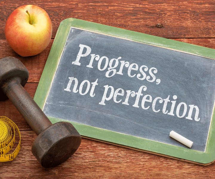an apple and a dumbell laying next to a chlkboard with progress not perfection written on it